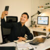 Mann der mit seinem Smartphone am Schreibtisch ein Selfie macht