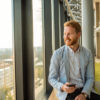 A smiling man in a blazer is sitting by the window of a modern office with a smartphone, looking out at the city.