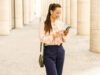 Eine lächelnde Frau mit Brille nutzt ihr Smartphone vor einer modernen Säulenarchitektur.