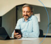 Ein lächelnder Mann im hellblauen Hemd sitzt am Schreibtisch und schaut auf sein Smartphone.