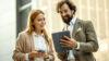 Eine Frau hat ein Handy in der Hand und schaut gemeinsam mit dem Mann, der neben ihr steht auf sein Tablet.